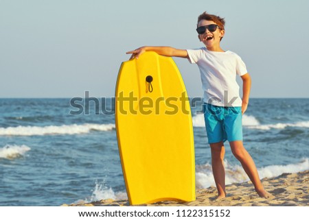 A happy boy in sunglasses in swimming trunks stands on the beach on golden sand and holds a swimming board, boogie board of yellow color against the blue sky and the sea with small waves in sunny sun