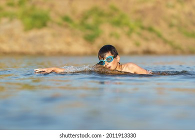 Happy Boy In Swimming Glasses Swims In The River In The Summer At Sunset. A Child Enjoys A Summer Children's Holiday On The Shore Of The Lake. Active Holidays. Dynamic Image