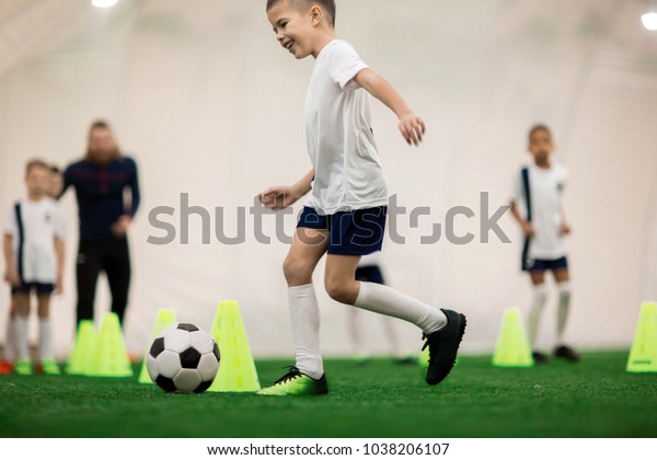 Happy boy in uniform kicking the ball while\
running around cones during\
training