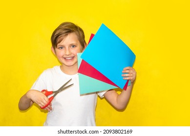 a happy boy in a white shirt holds scissors in one hand and colorful paper in the other hand on a yellow background