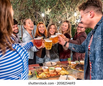 Happy Boys And Girls Toasting Beer In The Garden Of Restaurant - Group Of Friends Sitting On A Summer Terrace With Beer In Their Hands And Bbq On The Table - Friendship And Youth Concept