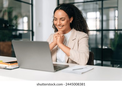 Happy brazilian or hispanic curly haired business woman, company ceo, recruitment manager, sitting at a work desk in a modern creative office, working in a laptop, studying information, smiling - Powered by Shutterstock