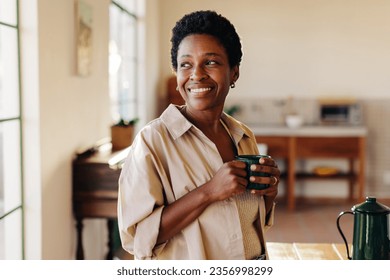 Happy Brazilian woman looking outside the window in her home kitchen, smiling and holding an enamel mug of plain coffee. Mature, afro-haired woman standing next to a breakfast table. - Powered by Shutterstock