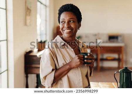 Happy Brazilian woman looking outside the window in her home kitchen, smiling and holding an enamel mug of plain coffee. Mature, afro-haired woman standing next to a breakfast table.