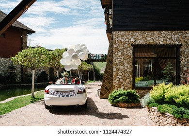 Happy Bride And Groom Arrived In A House Outside The City, Newlywed Wedding Couple Is Driving A Convertible Retro Car With Balloons On A Country Road For Honeymoon After The Ceremony. Just Married.