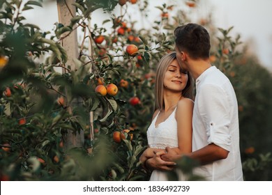 Happy bride and groom with closed eyes standing in embrace near trees in apple orchard. Man tenderly kissing him bride on the 
forehead.
