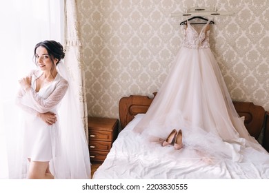 A Happy Bride In A Net Robe Is Standing By The Bed. A Wedding Dress And Shoes Are On The Hanger.