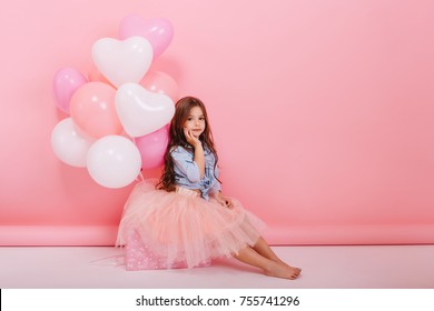 Happy brightful image of cute joyful little girl in tulle skirt sitting on present with balloons isolated on pink background. Amazing charming birthday fashionable kid looking to camera