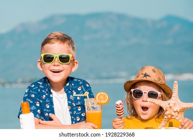 Happy Brother And Sister Eating An Ice Cream And Drinking Orange Juice On Sea Beach. Kids Boy And Girl In Sunglasses, Hat With Sea Star And Protection Sun Cream. Funny Summer Vacation For Family.