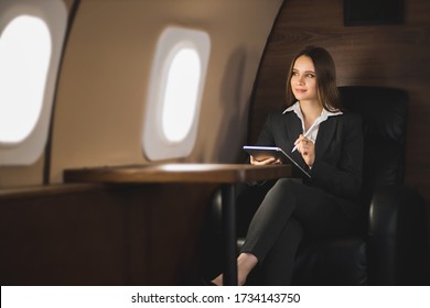 Happy Brown Haired Young Caucasian Businesswoman Holding Tablet, Sitting On Seat In Private Jet, Looking At Airplane Window And Smiling.