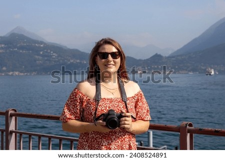 Happy brunette 30s woman enjoying summer holidays. Concept of vacation and travelling. Varenna, Italy. Lake Como. Redhead girl holding a camera outdoors at summer day
