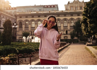 Happy brunette Asian woman in pink stylish hoodie and colorful sunglasses holds phone and listens to music in headphones outside.