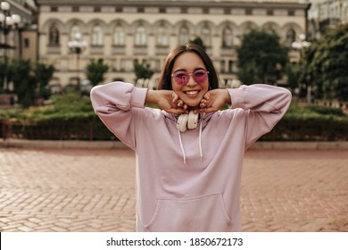 Happy brunette Asian woman smiles sincerely outside. Portrait of charming girl in pink hoodie and sunglasses. - Powered by Shutterstock