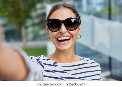 Happy brunette attraktive Frau trägt trendige Sonnenbrillen und gestreiftes Jumper hält Arm ausgestreckte Posen für Selbstvergnügen während der Freizeit im Freien steht auf unscharfem Hintergrund.