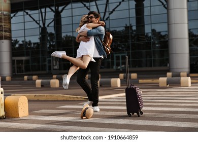 Happy Brunette Man Hugs His Girlfriend In White Dress. Woman Meets Boyfriend In Airport. Tourist Pose On Crosswalk With Suitcase.