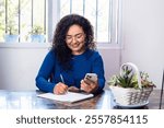 Happy brunette Mexican Latina woman working at home writing in notebook and holding cell phone