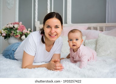 Happy Brunette Mom With Her Daughter Lies On The Bed In The Room And Looks At The Camera