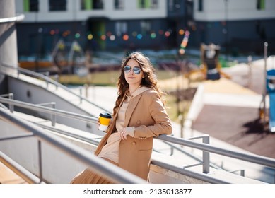 Happy Brunette Woman In Coat Enjoying A Walk Through The City On A Sunny Spring Day. A Woman Holds A Cup Of Coffee In His Hands.