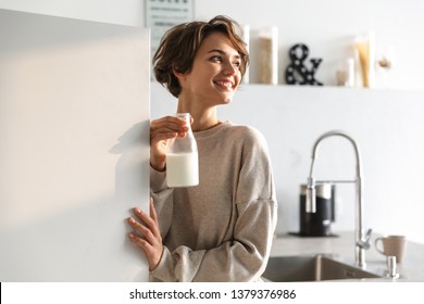 Happy brunette woman drinking milk and looking away while standing in kitchen