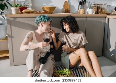 Happy brunette woman with glass of red wine putting piece of cheese in mouth of her girlfriend with myoelectric hand while sitting on the floor - Powered by Shutterstock