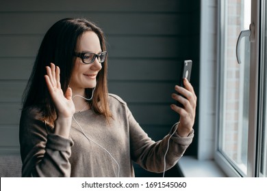 Happy brunette woman in glasses making facetime video calling with smartphone at home, using zoom meeting online app, social distancing, work from home, work remotely concept
