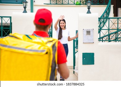 Happy Brunette Woman Greeting Deliveryman And Waving. Back View Of Deliveryman In Red Cap And Shirt Delivering Order At Home And Walking To Client. Delivery Service And Online Shopping Concept