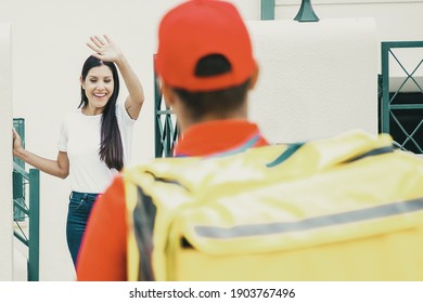 Happy Brunette Woman Greeting Deliveryman And Waving. Back View Of Deliveryman In Red Cap And Shirt Delivering Order At Home And Walking To Client. Delivery Service And Online Shopping Concept