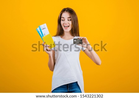 happy brunette woman with plane tickets in passport and credit card isolated over yellow