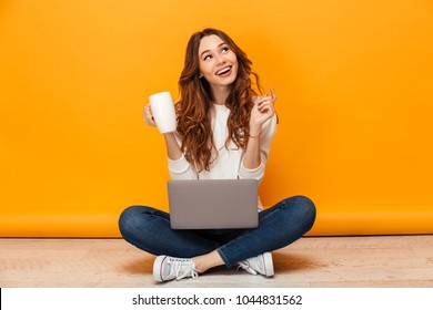 Happy Brunette Woman In Sweater Sitting On The Floor With Laptop Computer While Holding Cup Of Tea And Looking Away Over Yellow Background