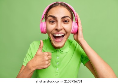 Happy Brunette Young European Woman Keeps Thumb Up Recommends Her Playlist Makes Approval Sign Wears Pink Stereo Headphones On Ears Dressed In Casual T Shirt Isolated Over Vivid Green Background.