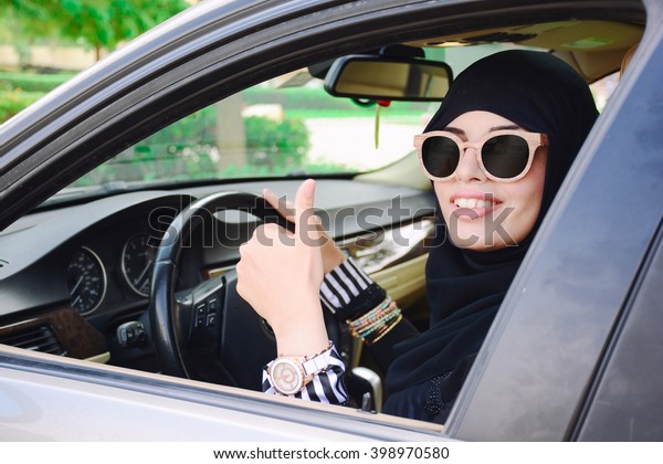 Happy business arabic lady\
in dark abaya and hijab driving her car and showing thumb up.\
Smiling arabian lady in sunglasses taking steering wheel of her new\
car.