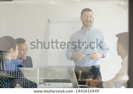 Happy business coach and multi ethnic team people laughing at funny joke at corporate training, cheerful diverse workers group with boss mentor manager having fun talking in boardroom behind glass