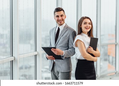 Happy business couple standing side by side, holding clipboards, looking at the camera, smiling.