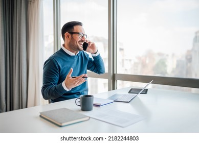 Happy Business Executive Discussing On Mobile Phone While Looking Away. Young Professional Is Wearing Smart Casuals. He Is Sitting At Desk In Corporate Office.