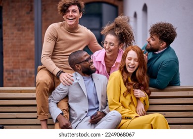 happy business executives talking in city park while taking coffee break on bench, having fun, laughing, talking jokes. attractive ladies and guys relaxing together, sitting on bench