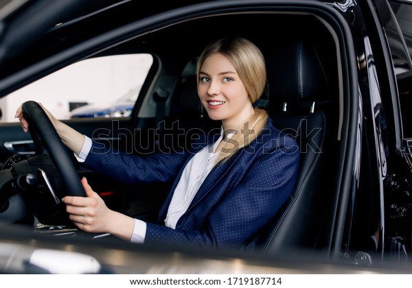 Happy business lady blonde in a blue suit sitting at\
the wheel of a new car