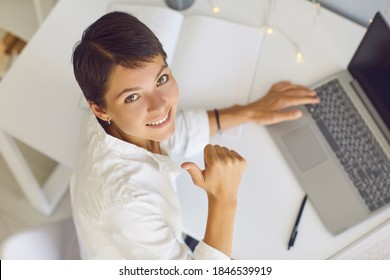 Happy business lady or freelancer looking up at camera, smiling and showing thumbs-up while working on laptop computer at home or in office. BYOD policy or remote work concept. From above, high angle