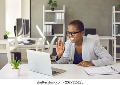 Happy Business Lady Having Virtual Online Video Call Meeting With Coworker. Black Woman In Suit Sitting At Office Desk, Looking At Laptop Screen, Smiling And Saying Hello Or Goodbye To Remote Employee