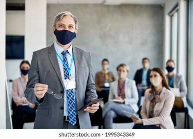 Happy Business Leader With Digital Tablet Standing In Board Room While Holding Seminar To Group Of Coworkers During COVID-19 Pandemic. 