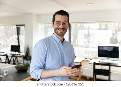 Happy Business Leader Wearing Glasses, Holding Cellphone, Looking At Camera. Portrait Of Businessman Posing In Office, Satisfied With Using Online App On Mobile Phone, Communication Concept