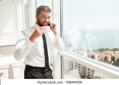 Happy Business Man Drinking Coffee And Talking By The Smartphone Near The Window