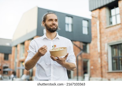 Happy Business Man Eating Healthy Salad On A Break Standing Over City Street Background. Male Dieting Nutrition Concept. Attractive Smiling Guy Enjoying Veggie Meal.