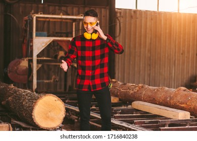 Happy Business Man In Protective Glasses And Headphones Talking Mobile Phone While Working At Timber Production Factory. Professional Sawmill Workman At Woodworking Warehouse. Small Business Owner