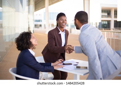 Happy business partners meeting in outdoor cafe and greeting each other. Business man and women standing and sitting at table outdoors, shaking hands, talking and smiling. Partners meeting concept - Powered by Shutterstock