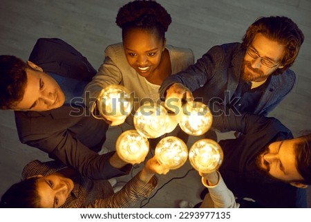 Happy business people lift up shining Edison light bulbs while standing together in office. Top view of multiracial people who have collective idea. Concept of ideas and innovations.