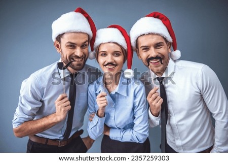 Happy business people in Santa hats are holding party props on sticks, looking at camera and smiling, on gray background