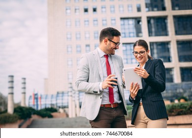 Happy Business People Working Outside On Tablet Computer In Front Of Tower Building.