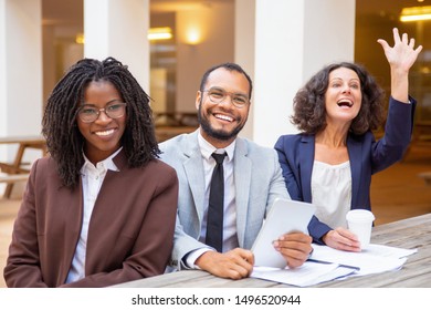 Happy Business Team Greeting Someone During Meeting, Training Or Conference. Business Man And Women Sitting At Table Outside, Waving Hello, Looking Into Distance And Smiling. Conference Concept