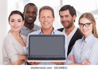 Happy Business Team. Group Of Cheerful Business People In Casual Wear Standing Close To Each Other While Mature Man Showing Laptop Monitor And Smiling