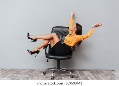 Happy Business Woman Sitting On Chair With Raised Arms And Having Fun Isolated Over Gray Background